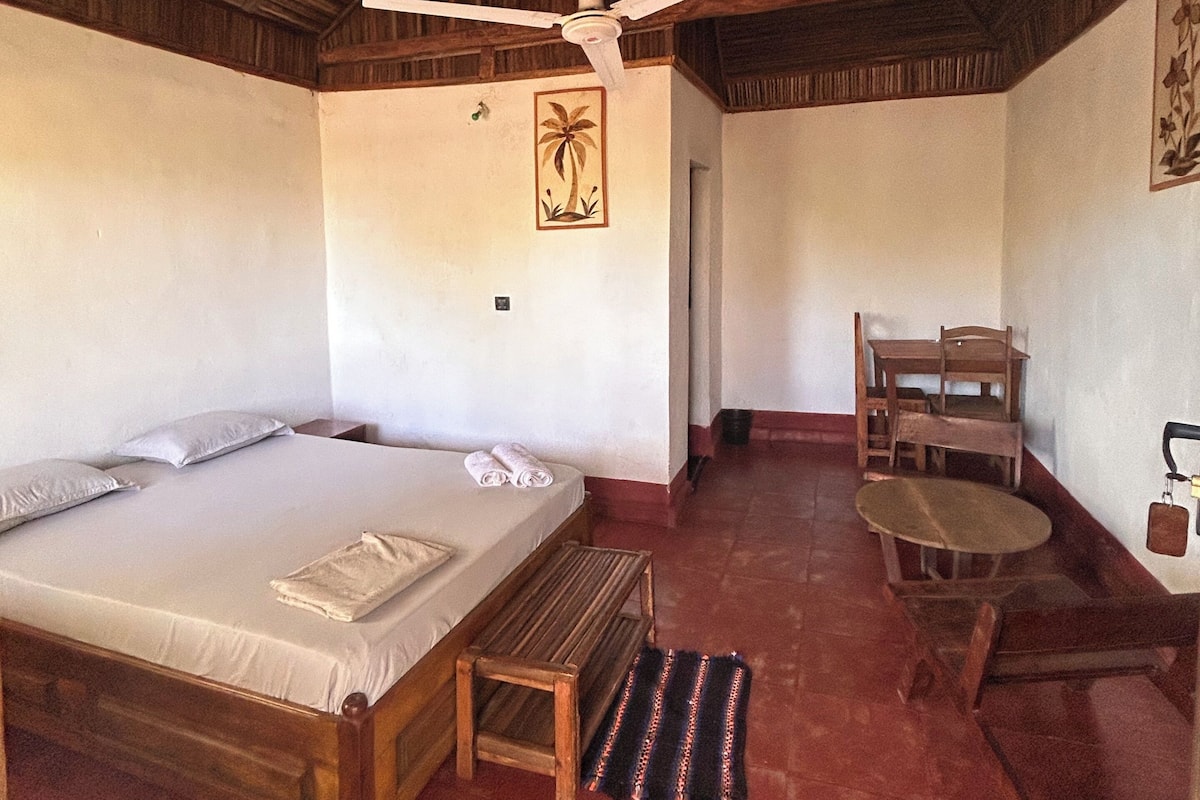 Une pièce comprenant un lit, une petite table, une chaise, un ventilateur de plafond et une porte donnant sur une autre pièce.