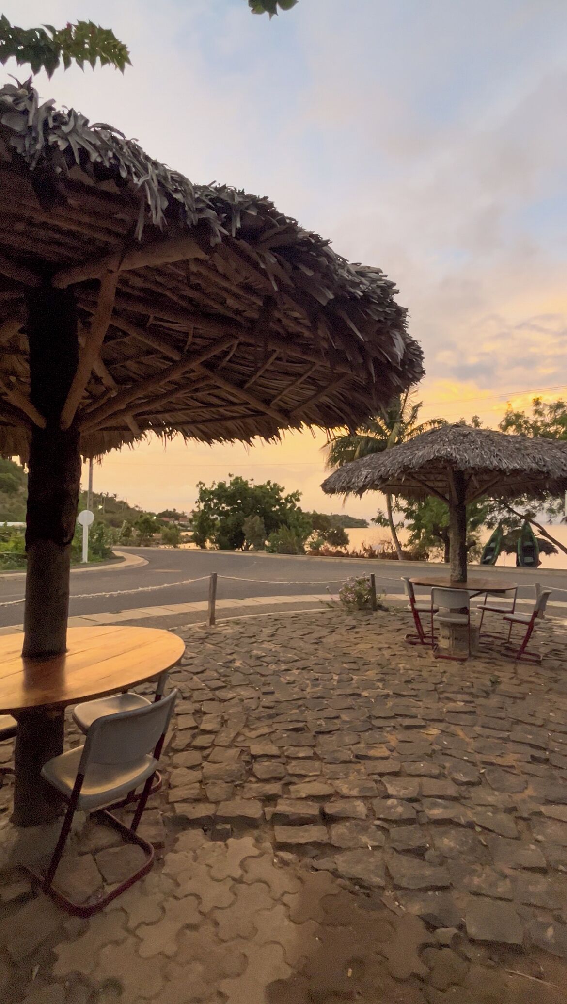 Un patio empedrado con una mesa y sillas de madera, rodeado de sombrillas de paja, y una carretera al fondo.