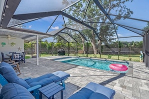 A covered outdoor swimming pool with lounge chairs, a table, and a grill area.