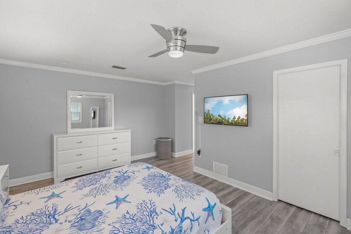 A bedroom with a bed, dresser, ceiling fan, and a wall-mounted TV.