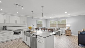 Une cuisine moderne avec des meubles blancs, un îlot central et un coin repas comprenant un canapé gris et des tables d’appoint en bois.