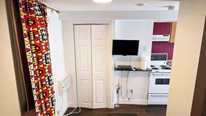 Uma cozinha compacta com eletrodomésticos brancos, uma TV de tela plana acima do fogão e uma porta que dá para outro cômodo.