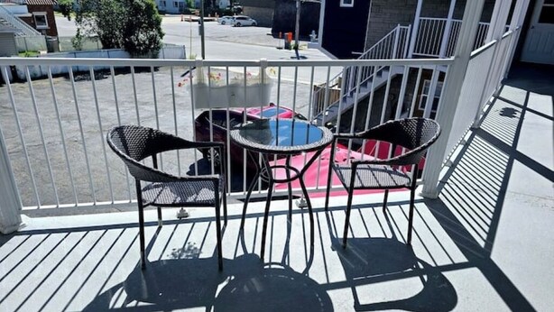 Varanda com duas cadeiras e uma mesa pequena, com vista para uma rua onde se vê um carro vermelho e um edifício ao fundo.