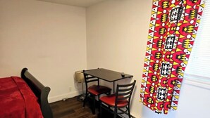 Um quarto pequeno com uma mesa de madeira e duas cadeiras, um cobertor vermelho em uma cadeira, e uma cortina estampada na janela.