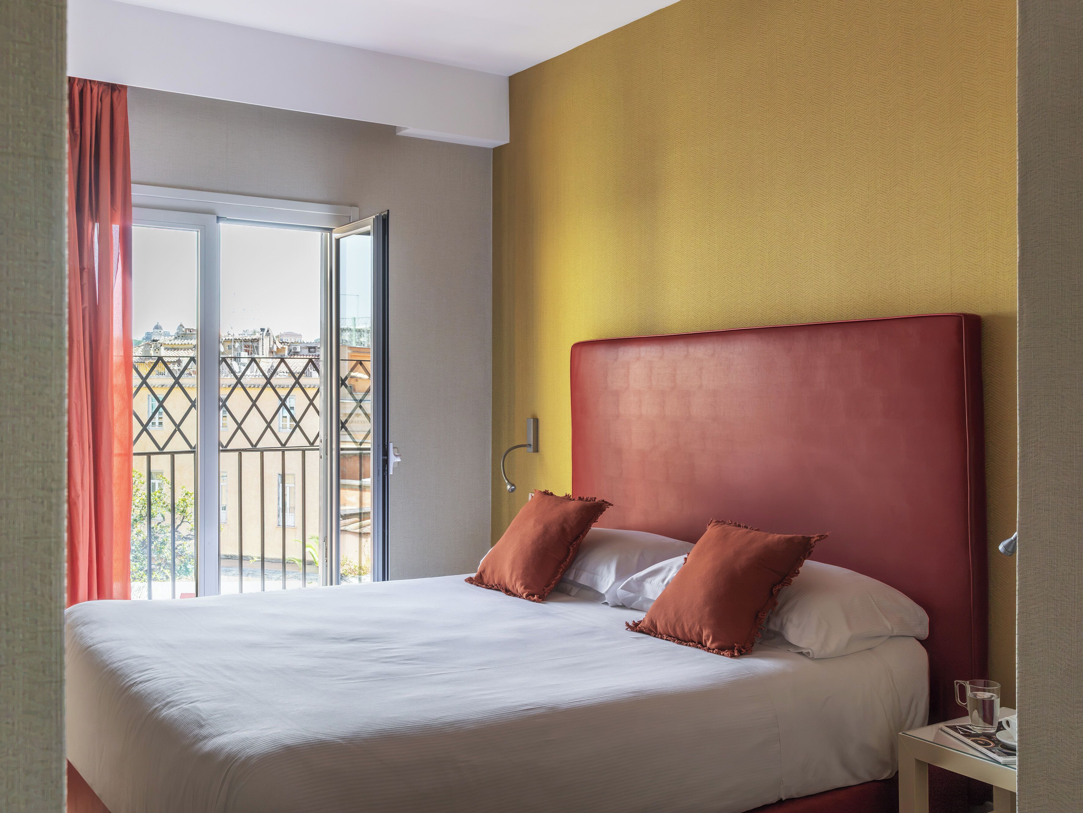 A hotel room with a bed, a nightstand, a window with curtains, and a view of buildings outside.