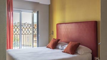 A hotel room with a bed, a nightstand, a window with curtains, and a view of buildings outside.