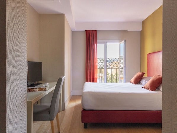 Quarto de hotel com cama, escrivaninha, cadeira, televisão e janela com vista para edifícios.