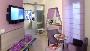 Un salon moderne avec un téléviseur à écran plat, des meubles violets, un magazine posé sur une table d’appoint et un petit coin cuisine à l’arrière-plan.