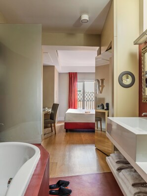 Une chambre d’hôtel avec un grand lit, un bureau, une chaise, une salle de bain avec baignoire et lavabo, et une vue sur l’extérieur.