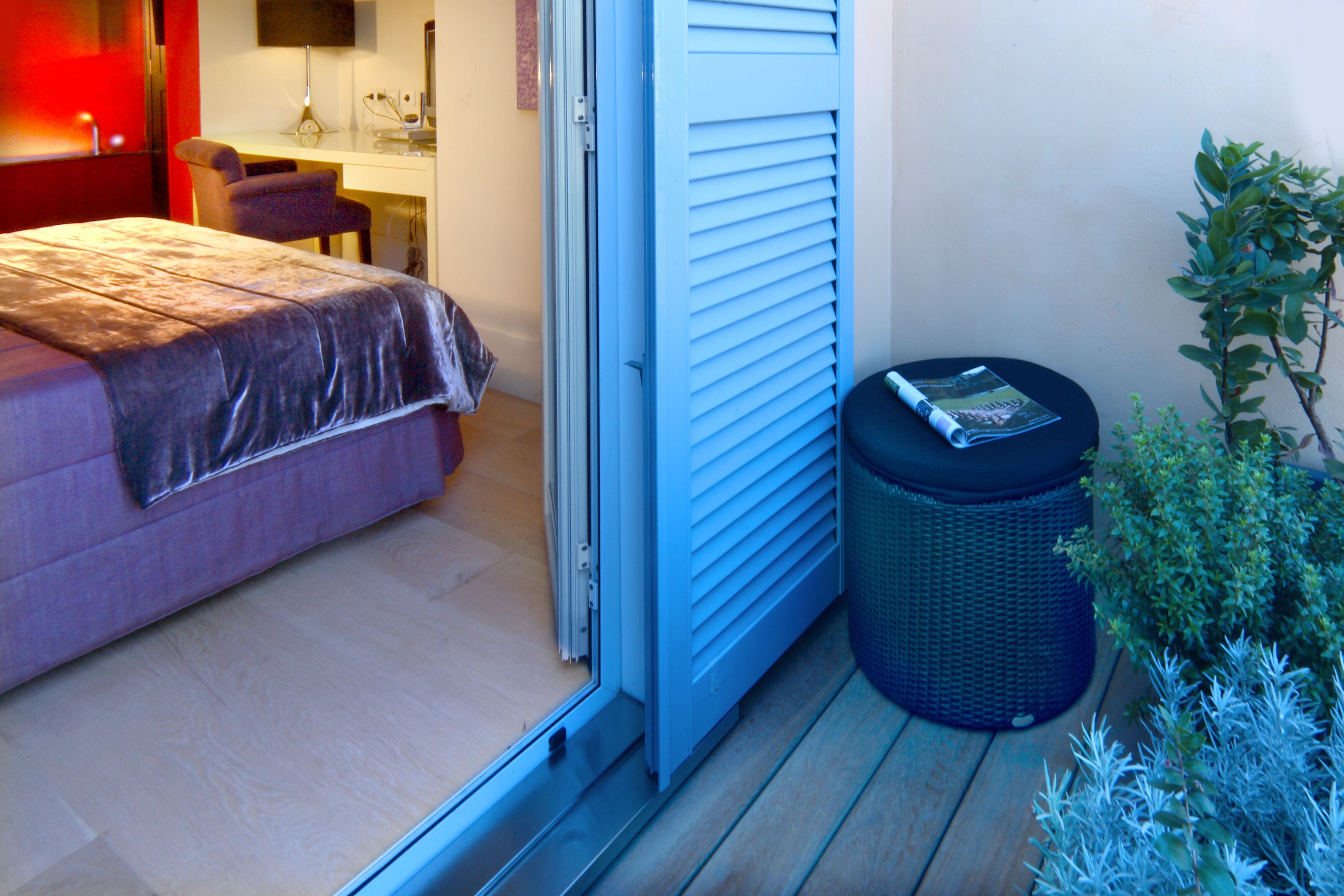 A bedroom with a bed, a desk, and a chair. There is a window with blinds, a trash bin, and a small plant.