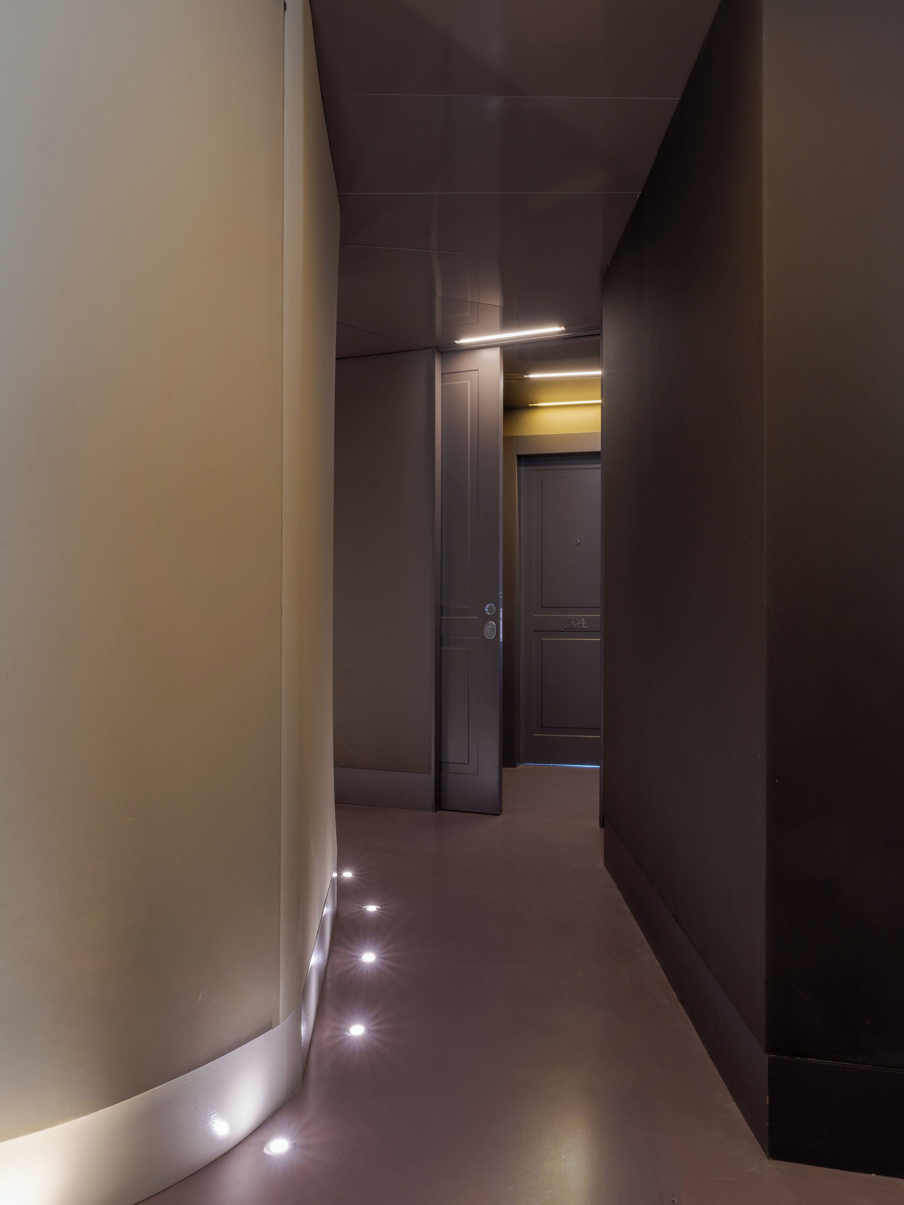 A hallway with a door on the right, a light strip on the wall, and a reflective floor.