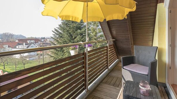 Un balcón con un paraguas amarillo, mobiliario de mimbre y vistas a casas y árboles.