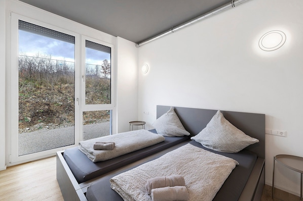 A modern bedroom with a large sliding glass door, a bed with grey bedding, and a small bedside table.