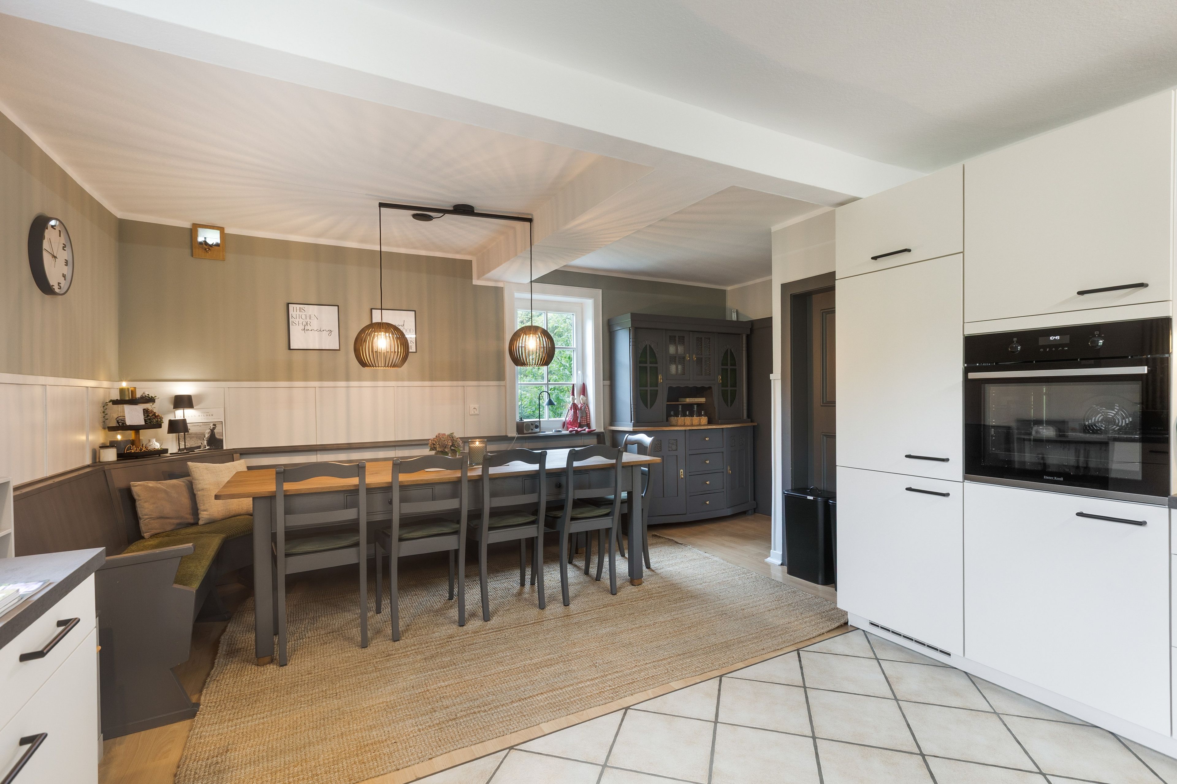 A modern kitchen with a dining area, featuring a built-in oven, a large island with seating, and a clock on the wall.