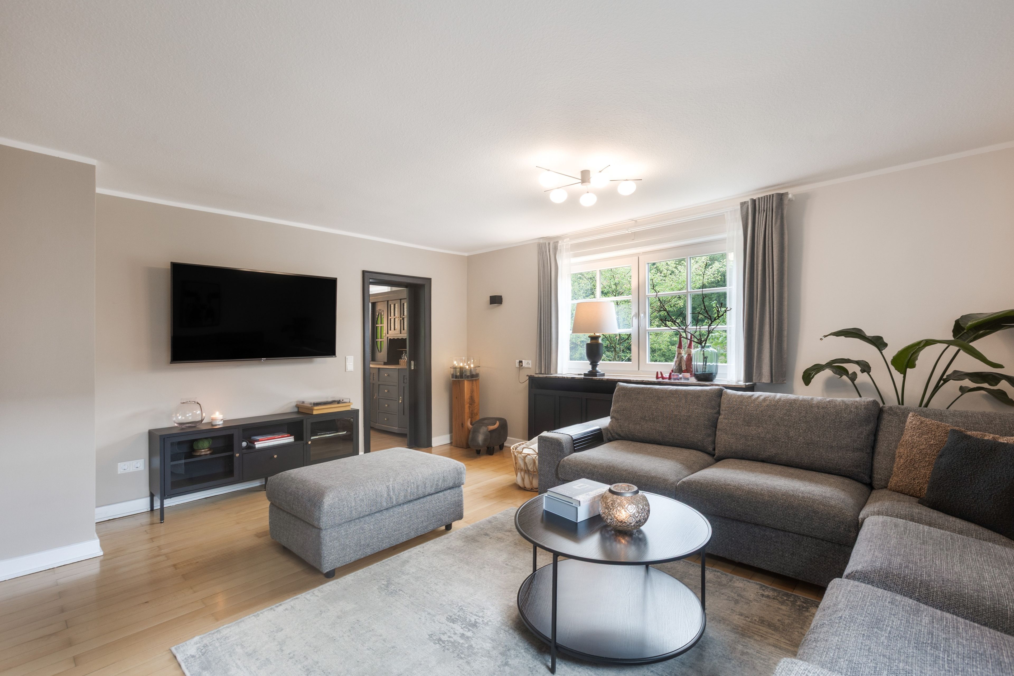 Een moderne woonkamer met een flatscreen televisie, grijze banken, een salontafel en een groot raam met gordijnen.
