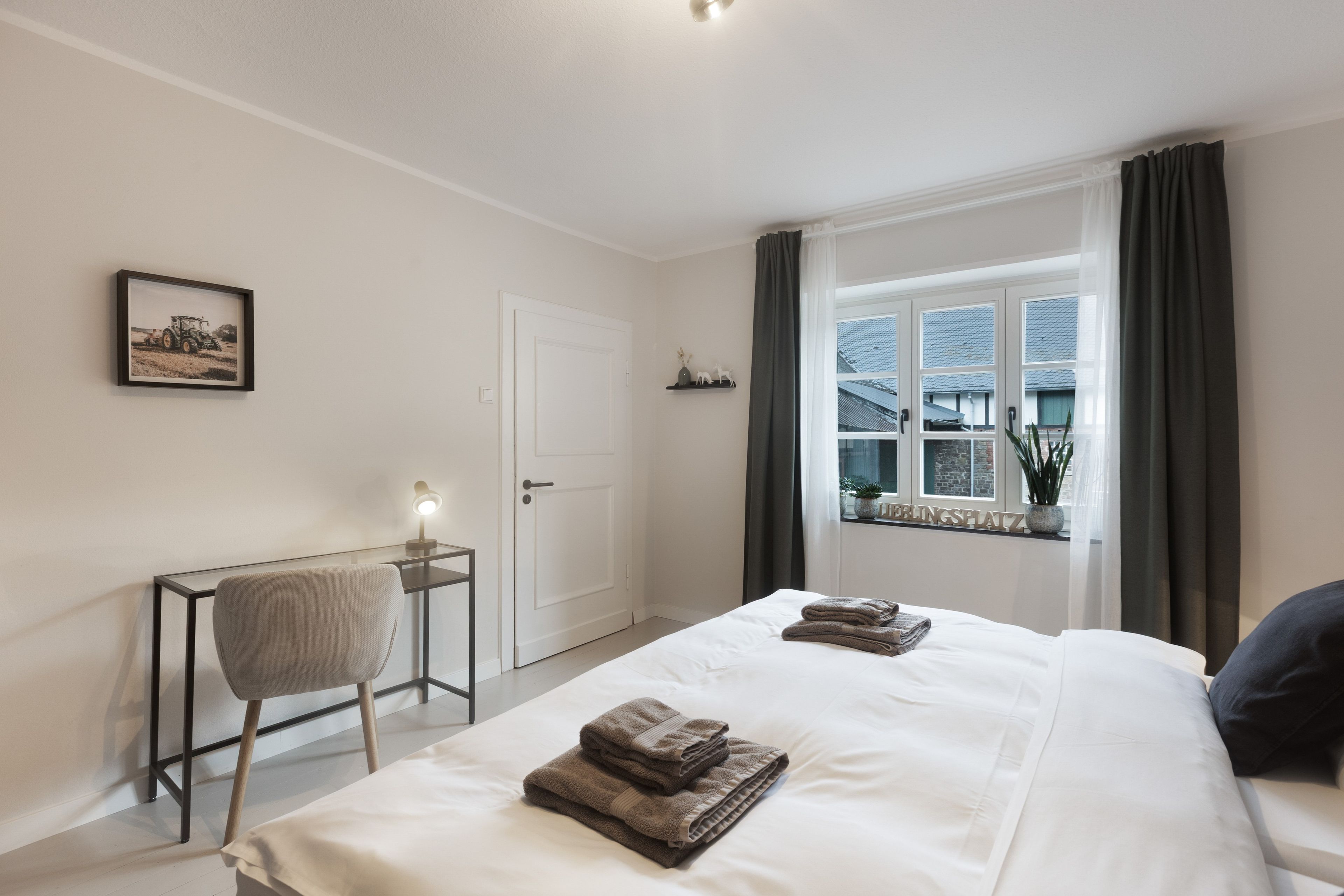 A hotel room with a bed, a desk with a chair, a window with curtains, and a framed picture on the wall.