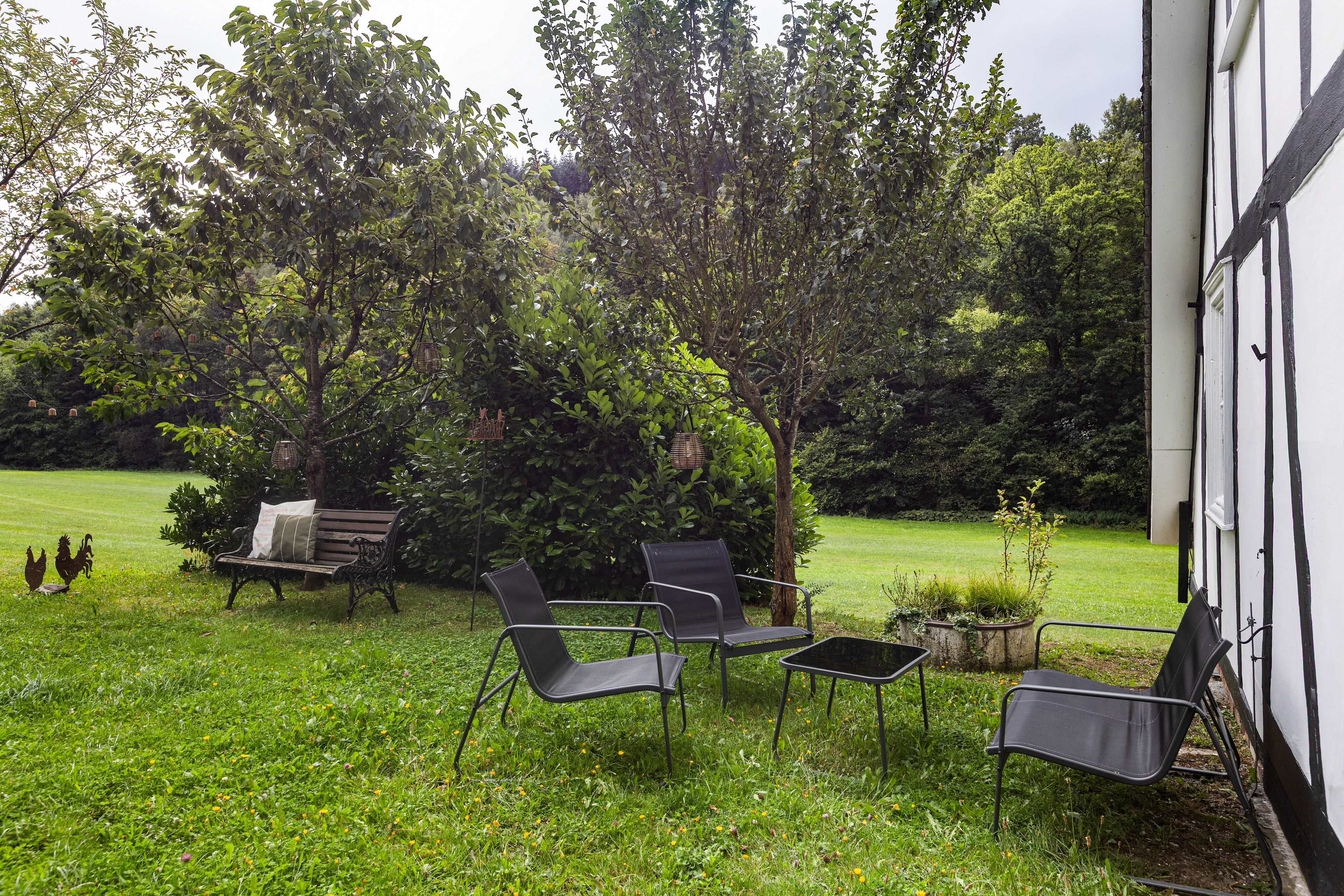 An outdoor seating area with metal chairs and a bench, surrounded by lush greenery and trees.