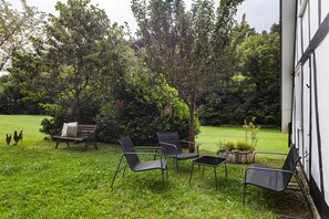 Un área de estar al aire libre con sillas de metal y un banco, rodeada de abundante vegetación y árboles.