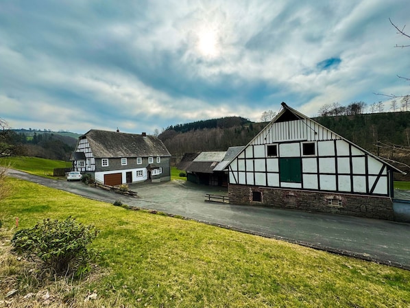 A traditional half-timbered building with a stone foundation, a car parked in front, and a grassy area with a path leading to it.