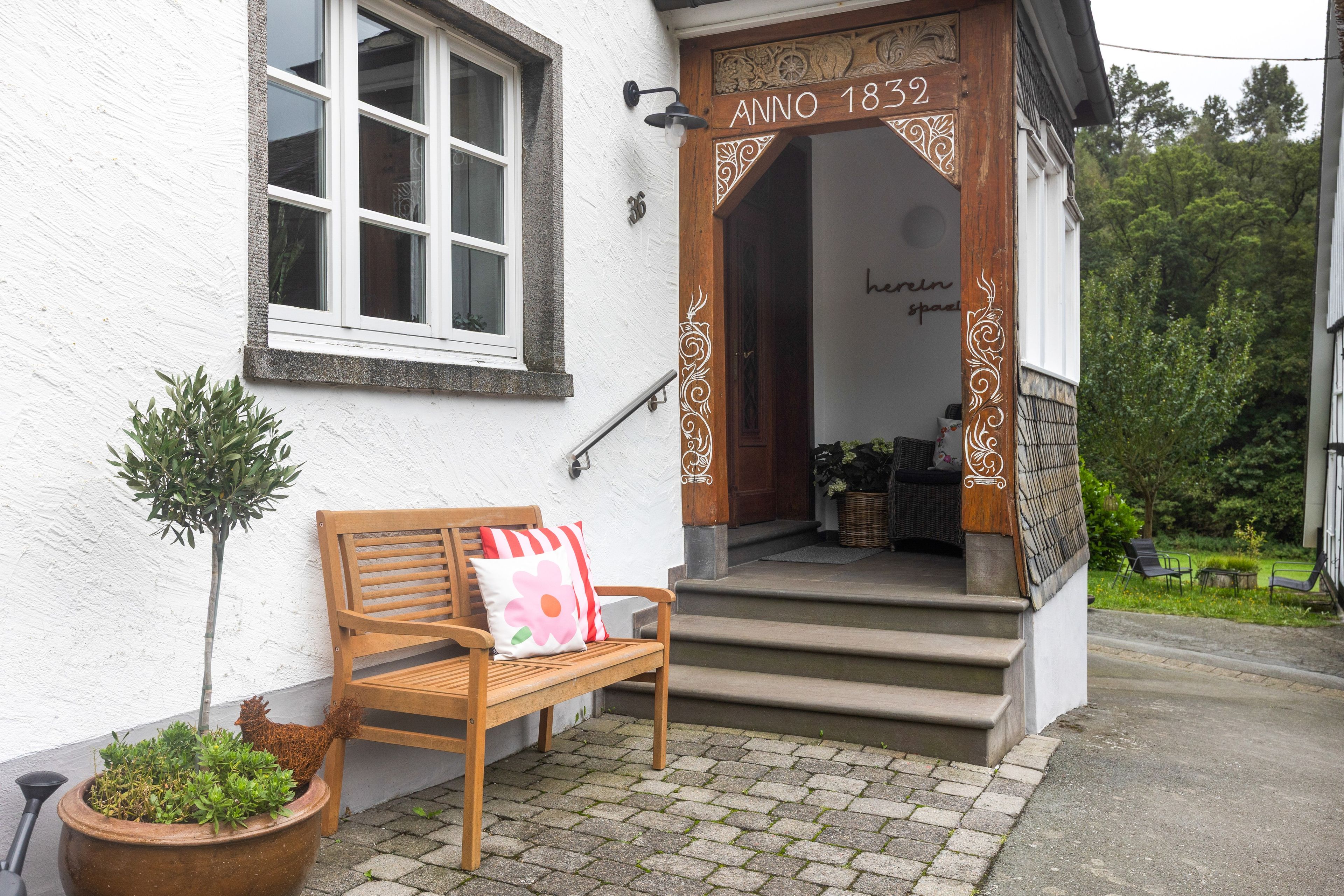 A wooden bench with a cushion outside a building with a white exterior and a wooden door marked 'Anno 1832'.