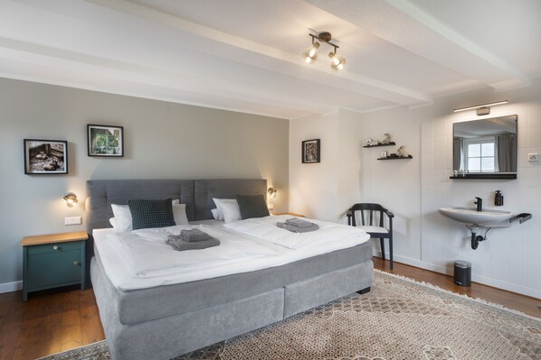 A modern bedroom with a large bed, bedside tables, a chair, a bathroom with a sink and mirror, and wall-mounted lighting.