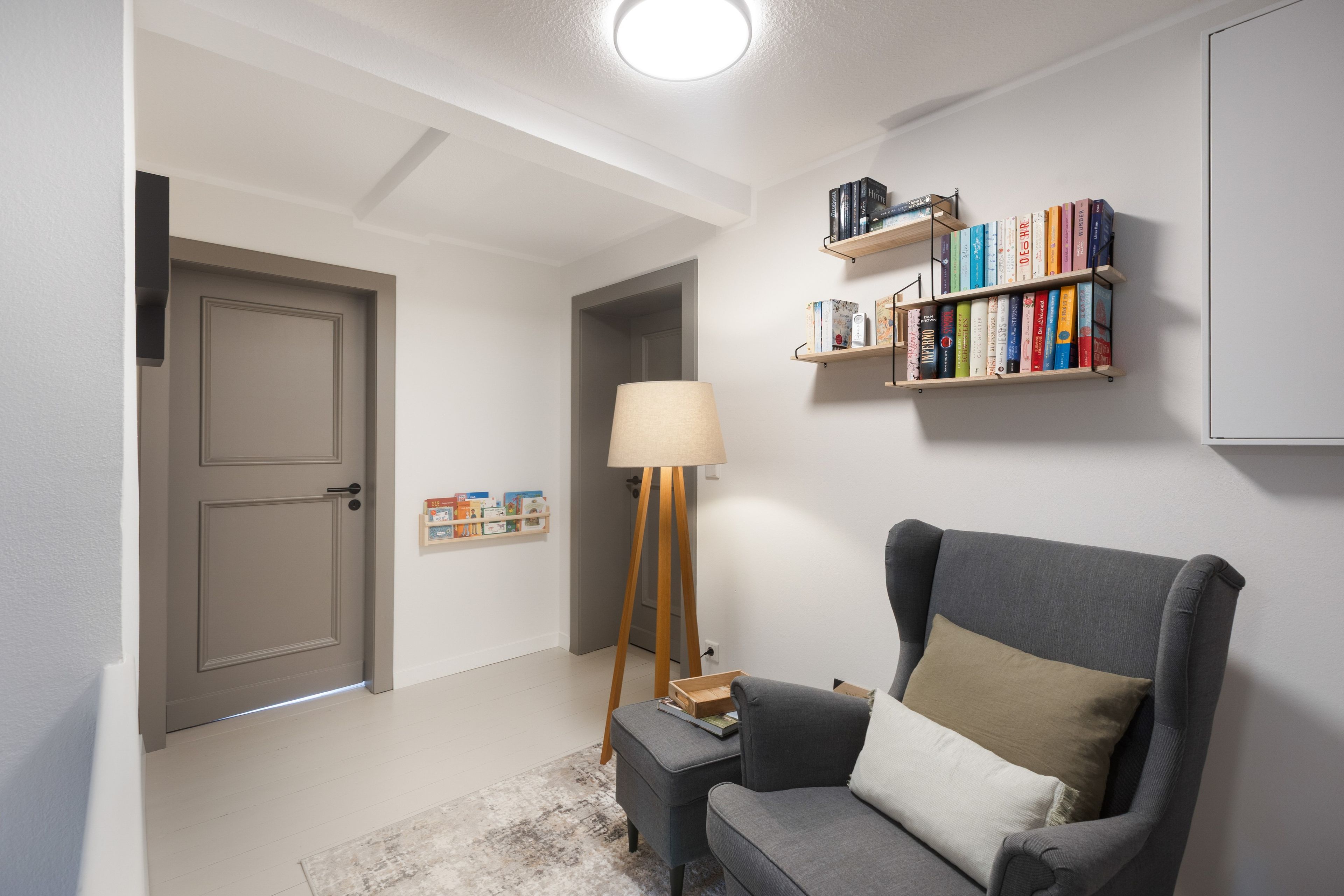 A room with a grey armchair, a floor lamp, a bookshelf, and a door.