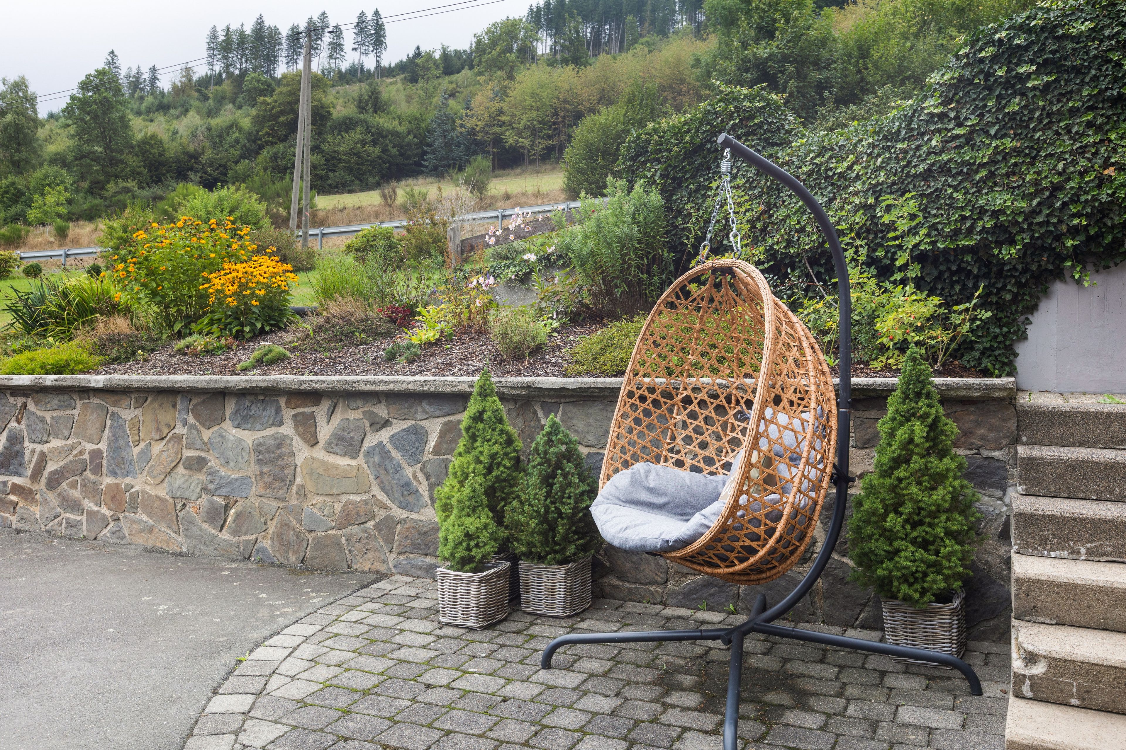 Uma cadeira suspensa de vime com almofada, colocada em uma parede de pedra com vista para o jardim.