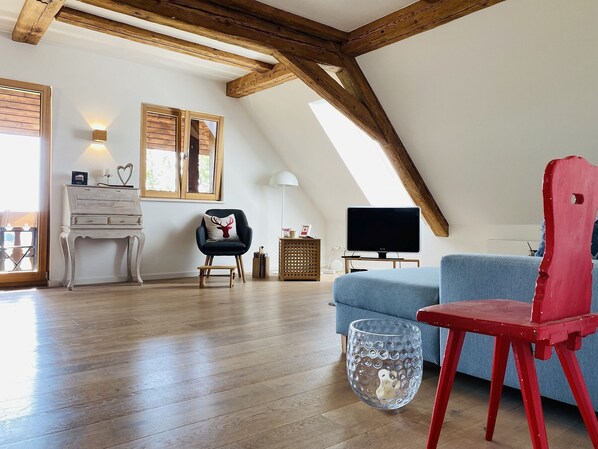 Une pièce de style loft avec des poutres en bois, une télévision, un canapé bleu, un fauteuil rouge et une petite table avec une lampe.