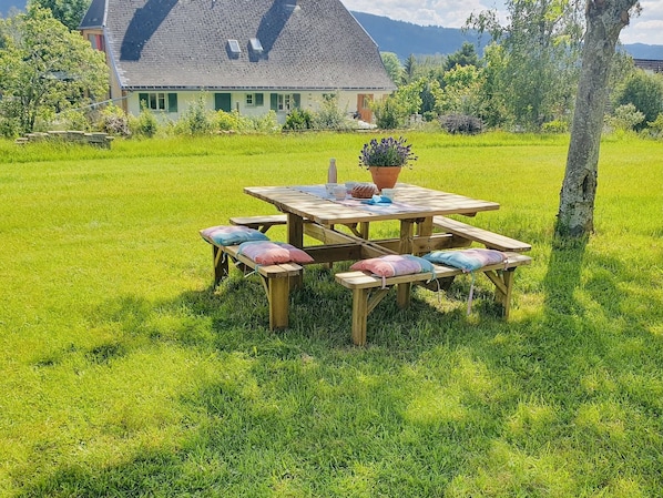 A picnic table with benches and cushions in a grassy field with a house in the background.