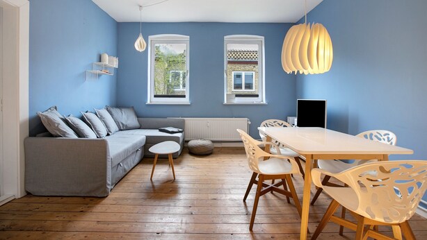 Ein Zimmer mit blauer Wand, einem grauen Sofa, einem Holzesstisch mit Stühlen und einer hängenden Leuchte.