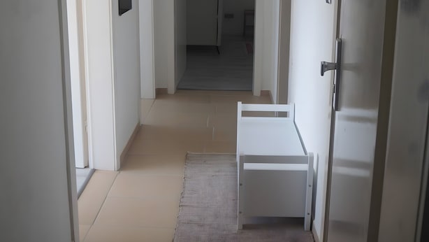 En smal hall med vita väggar, ett kaklat golv och en dörr på höger sida.