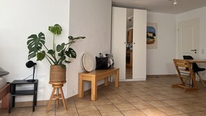 Uma sala de estar moderna com uma planta grande, um banco de madeira, uma mesa de jantar e uma cozinha visível por uma porta aberta.
