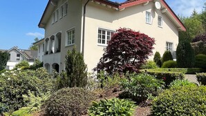 Um edifício de dois andares com telhado de telhas vermelhas, várias janelas e um jardim bem cuidado com diversas plantas e arbustos.