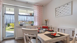 Un comedor con una mesa dispuesta para dos, sillas blancas, un radiador y una ventana con cortinas.