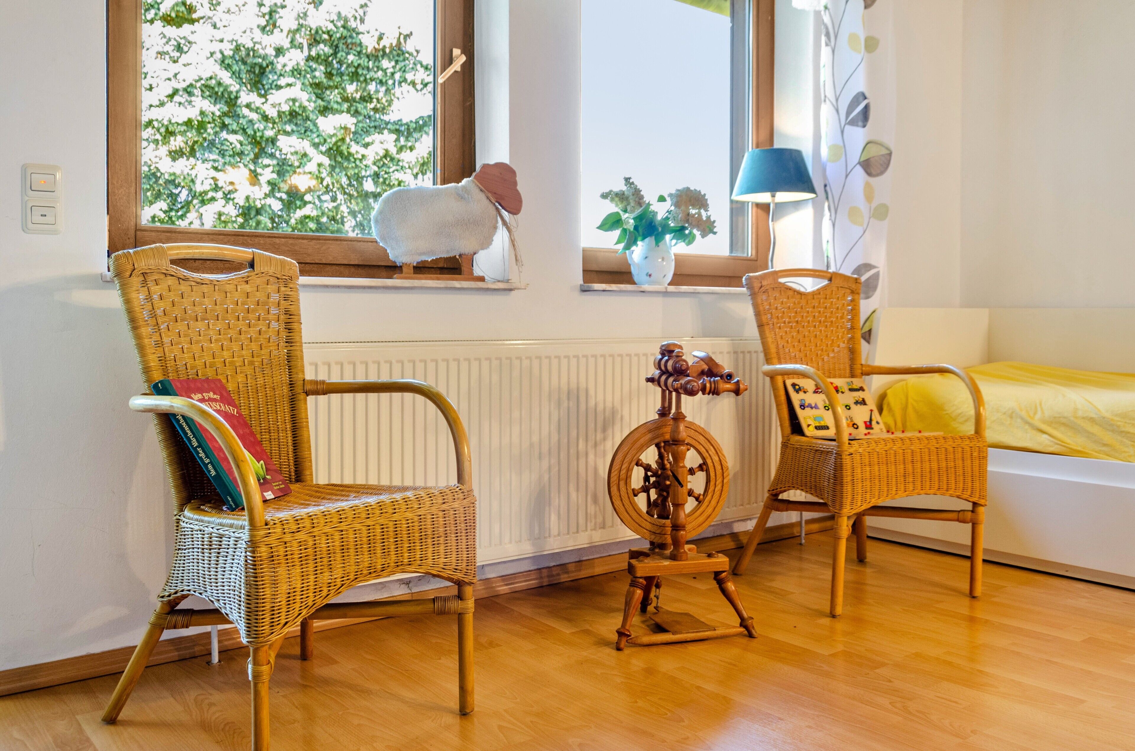 Une pièce avec deux fauteuils en osier, une petite table sur laquelle se trouve un rouet, une fenêtre donnant sur des arbres et un lit recouvert d’une couverture jaune.