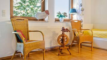 Une pièce avec deux fauteuils en osier, une petite table sur laquelle se trouve un rouet, une fenêtre donnant sur des arbres et un lit recouvert d’une couverture jaune.