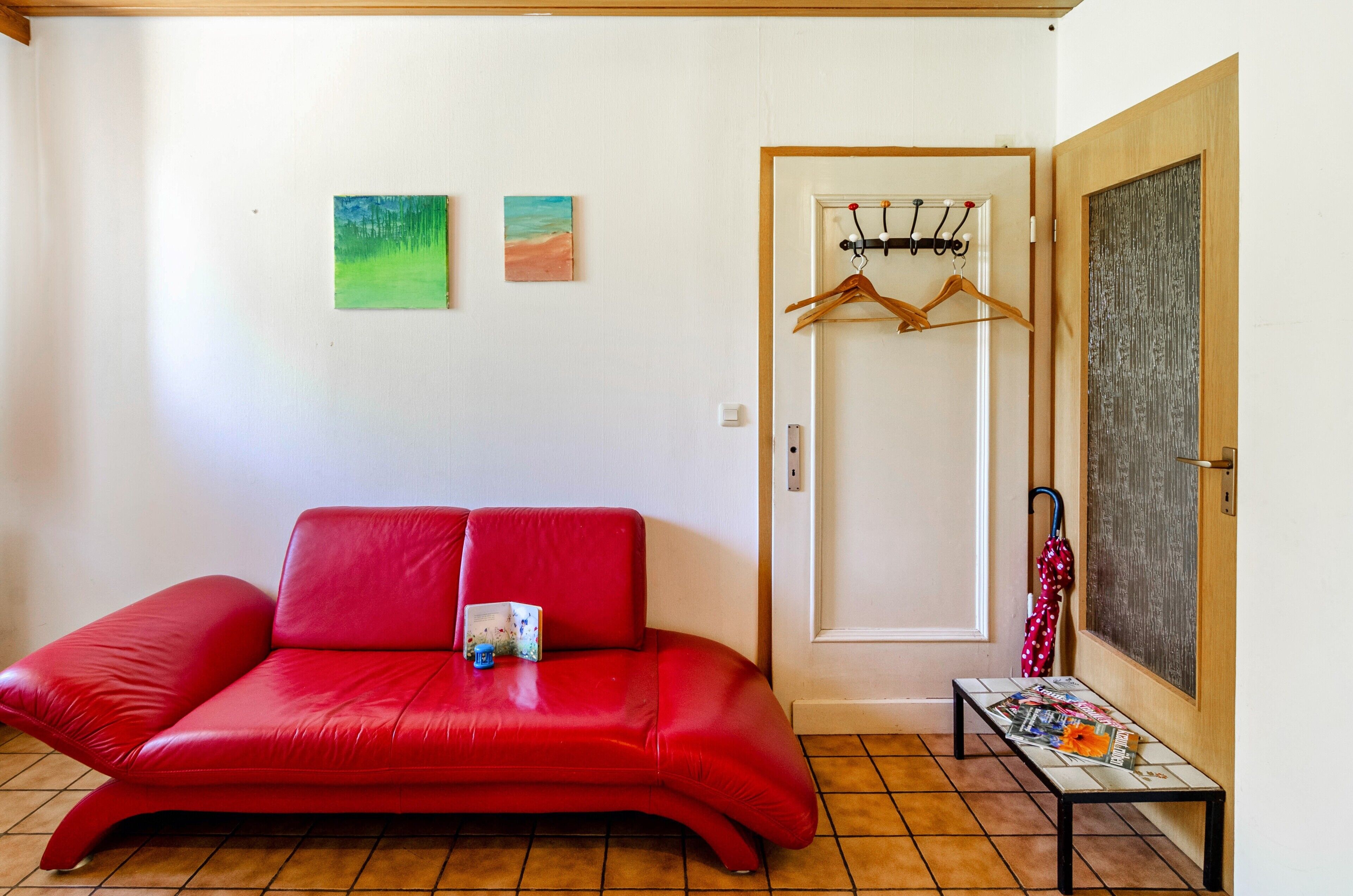 A red sofa, a bench with magazines, a door with a coat rack, and two paintings on the wall.