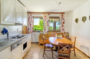 A kitchen with white cabinets, a wooden dining table, and a view of greenery outside. - Apartment 'Landleben' with Mountain View and Shared Garden (Hofgeismar)