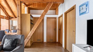 Eine Holztreppe führt zu den oberen Stockwerken, ein graues Sofa mit Kissen, eine weiße TV-Konsole und Holztüren.