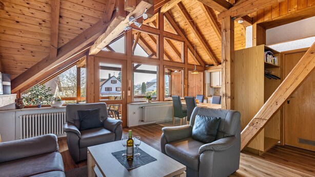 Ein geräumiger Loft-Raum mit hohen Decken, Holzbalken und großen Fenstern. Es gibt Ledersofas, einen Holztisch und eine Treppe, die zu einer erhöhten Ebene führt.