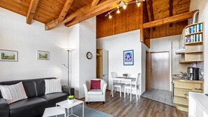 Una sala de estar moderna con un sofá negro, sillones blancos, una mesa de comedor y una zona de cocina con armarios de madera y una estantería empotrada.