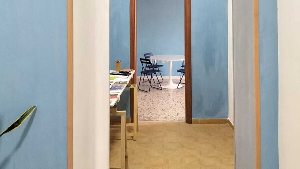 A narrow hallway with a blue wall, a wooden door frame, and a small table with chairs in the distance. - Art house - Central station (Napoli)