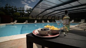 Mesa junto a la piscina con una botella de vino blanco, dos copas, fuentes de aperitivos y una piscina al fondo.