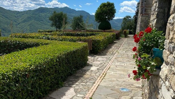 Un sendero de piedra serpentea por un jardín cuidado, con setos y plantas florecientes, bordeado por muros de piedra y un paisaje montañoso de fondo.