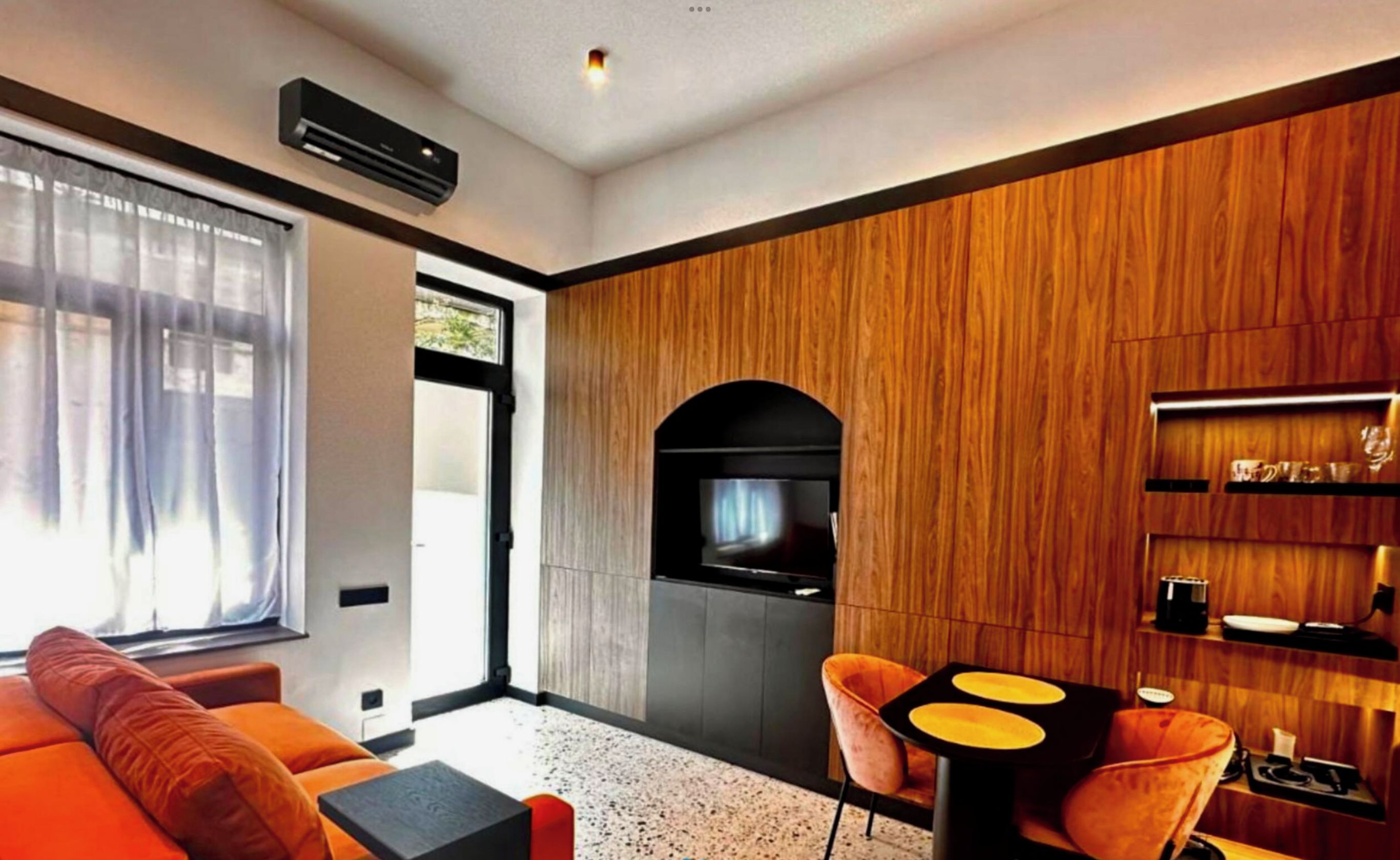 Una sala moderna con un sofá, una mesa de centro y un televisor. Se aprecia una pared de madera y una zona de cocina con un microondas.