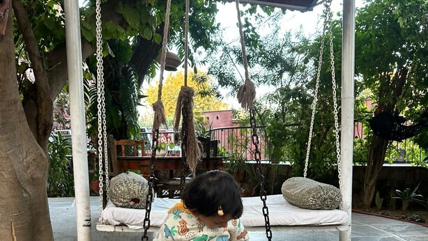 A child on a swing with a patterned seat, surrounded by trees and a covered area with a tiled floor. - VISHAL VILLA  (Jaipur)