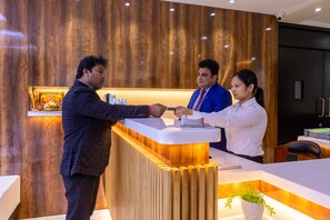 A reception desk with a wooden panel backdrop, a white counter, and a potted plant. - The SM Hotel By RMR Hotels (New Delhi)