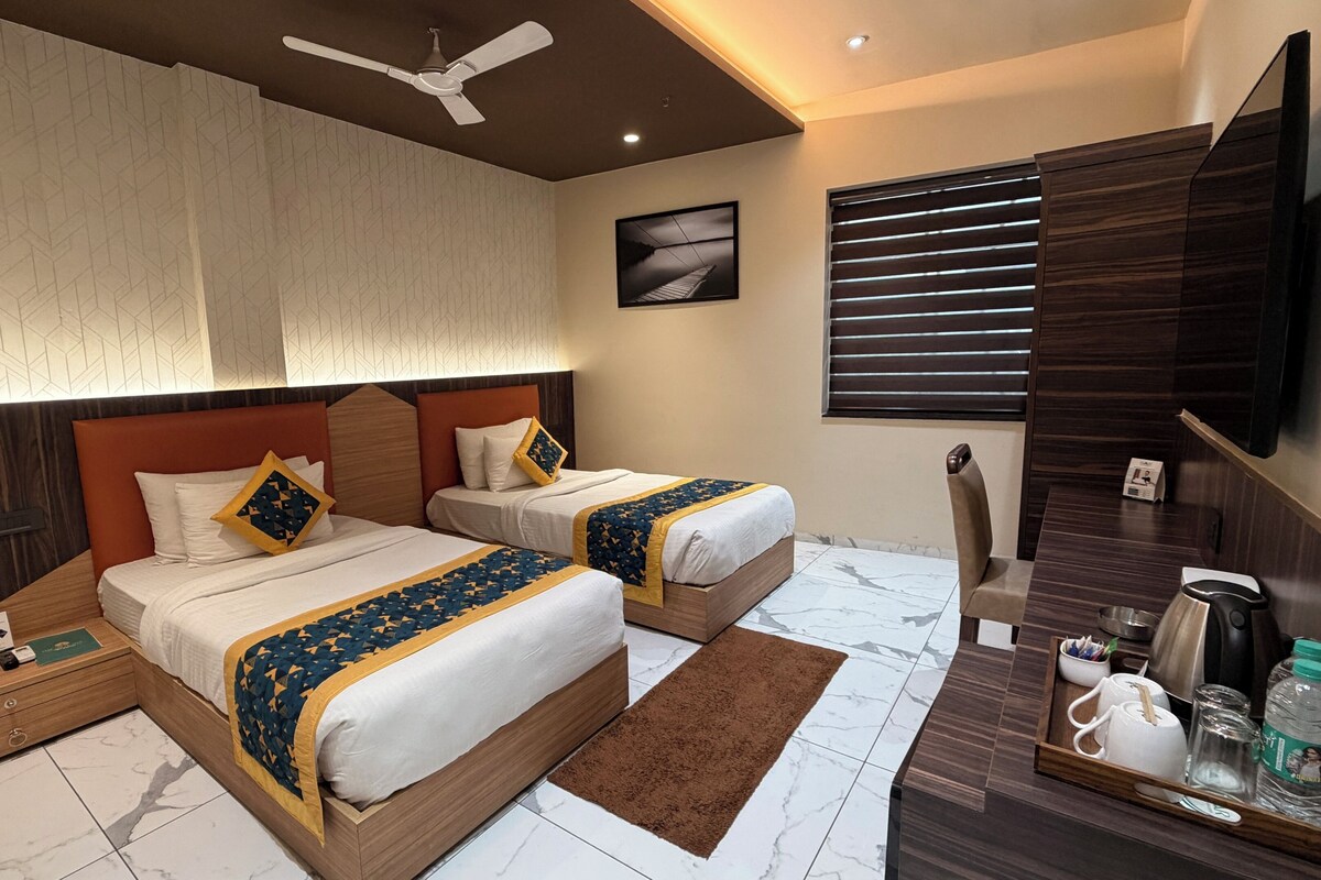 A hotel room with two beds, a desk, a coffee station, and a large window with blinds.
