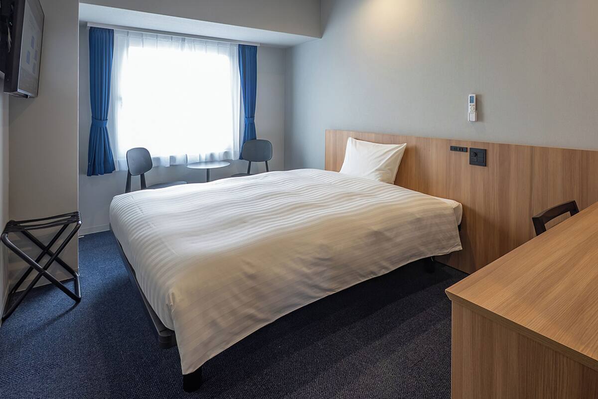 Quarto de hotel com uma cama grande, uma escrivaninha, uma cadeira, uma televisão e uma janela com cortinas.