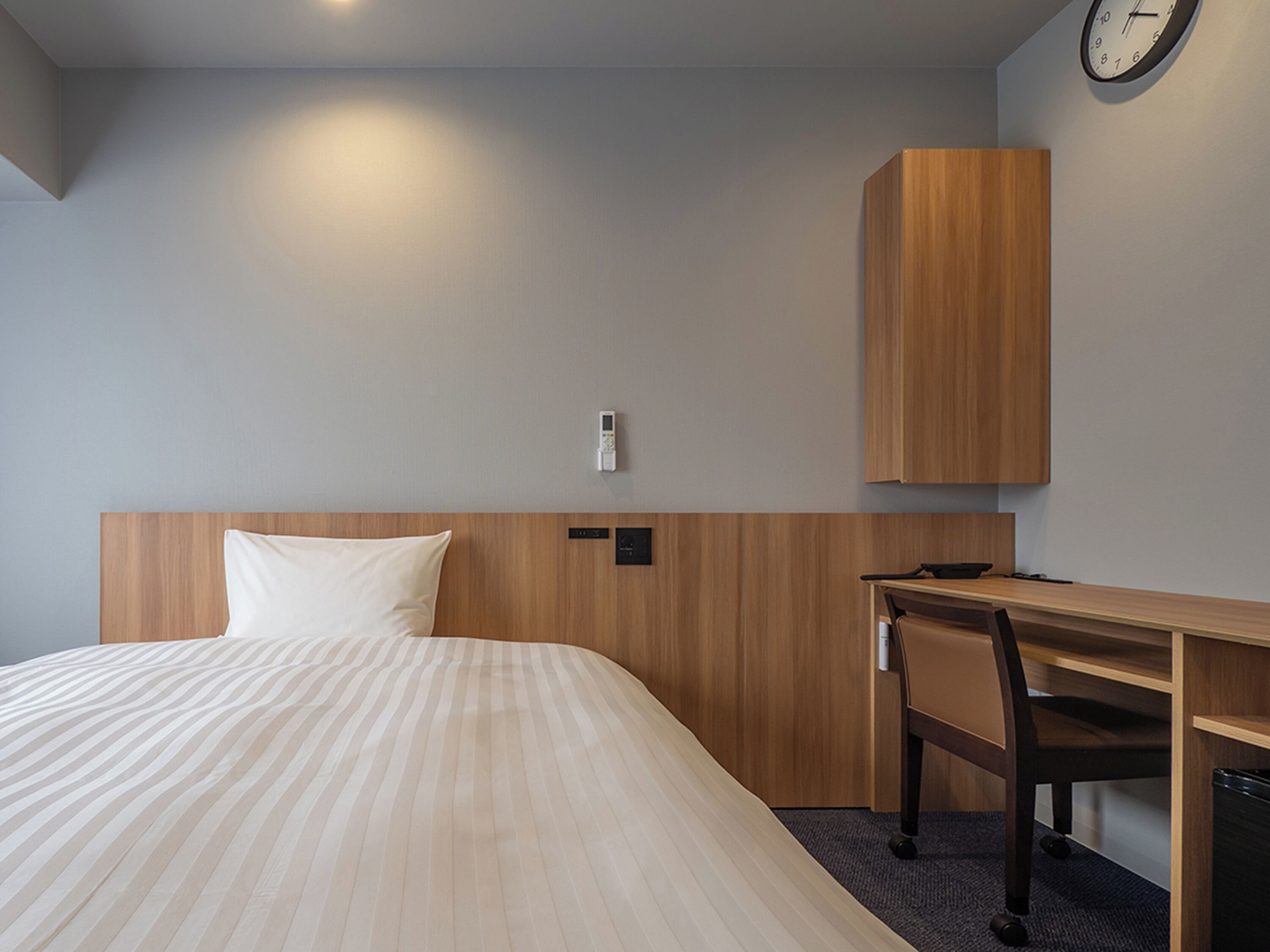 Quarto de hotel com uma cama grande, uma escrivaninha de madeira, uma cadeira, um relógio na parede e um termostato.
